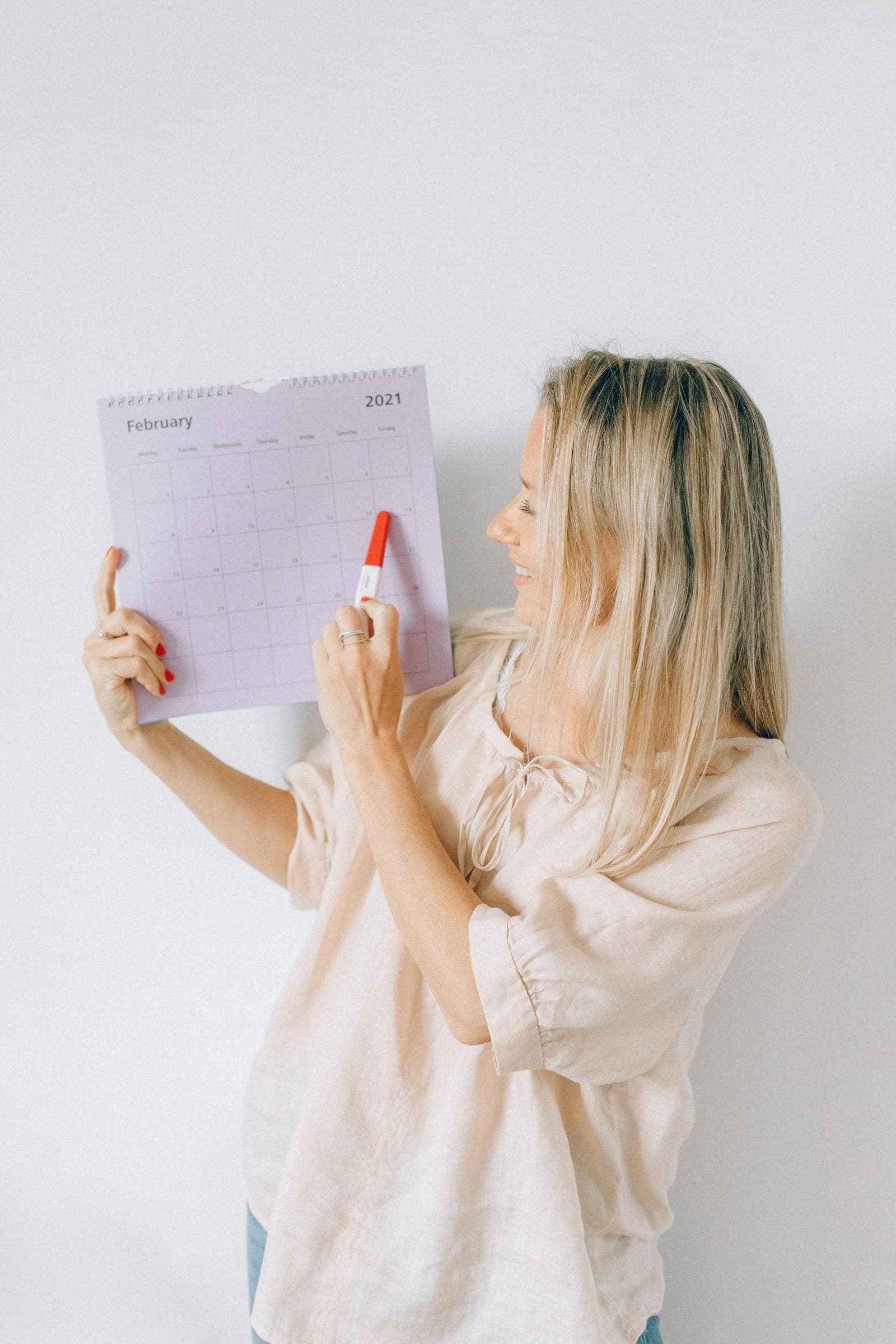 A woman holding up a wall calendar and pointing at a date, marking a recurring monthly appointment.