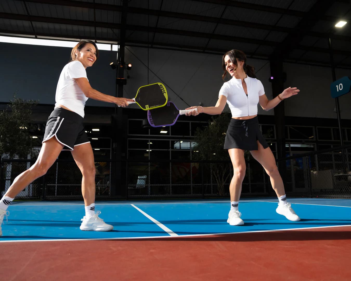 Two women laughing and playing pickleball on an indoor blue court, paddles raised.