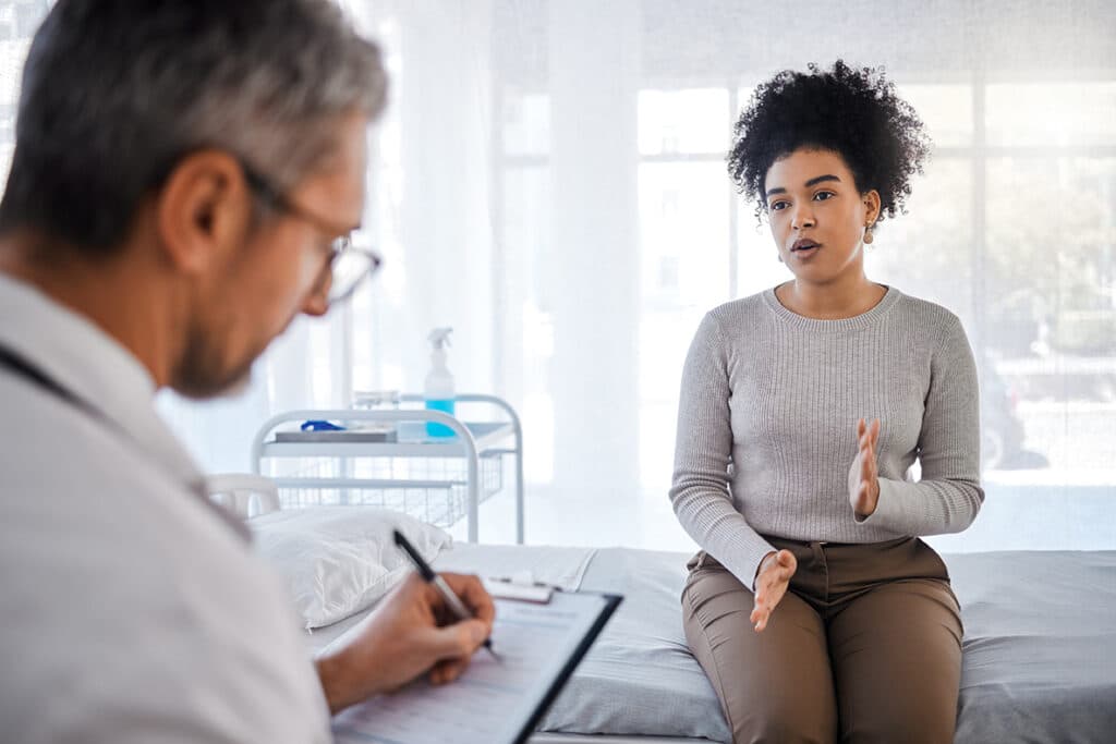 A doctor writing notes on a clipboard while a patient explains her symptoms during a clinic consultation.