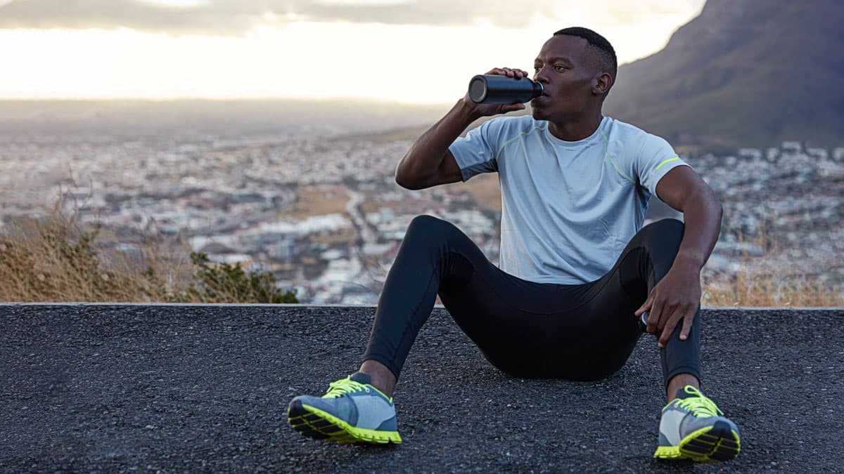 A runner sitting and drinking from a water bottle overlooking a city skyline after a run.